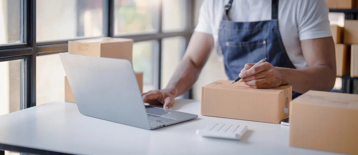 A small-business owner prepares online orders beside a laptop and packing boxes.